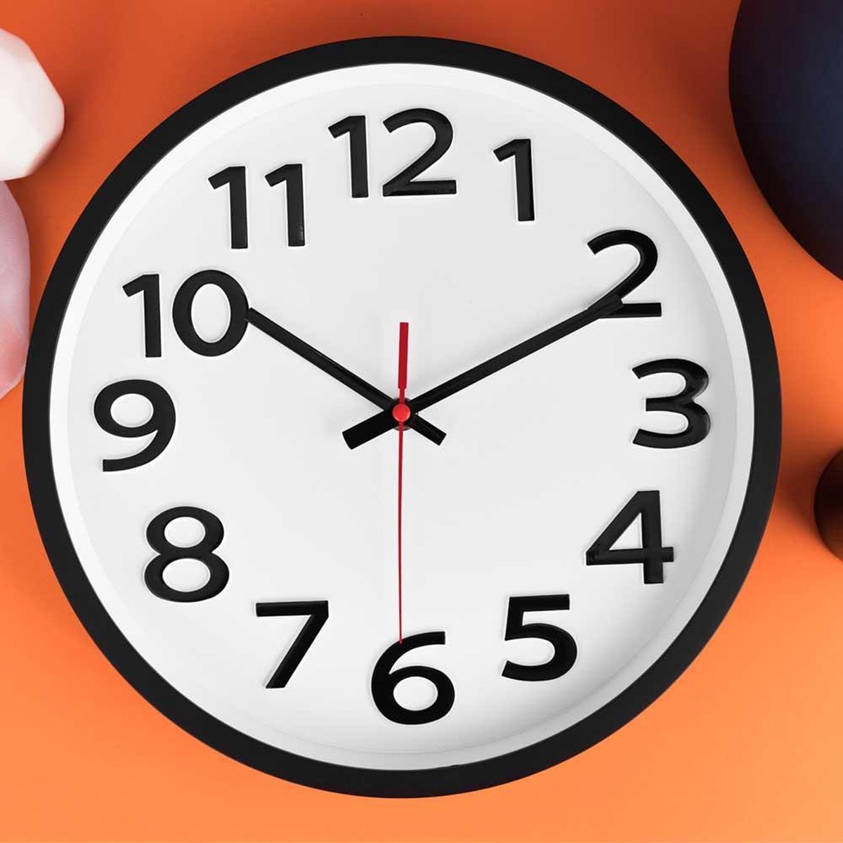 Round white clock face close-up on orange surface, bold raised black numerals, black hands, red second hand, hand visible…
