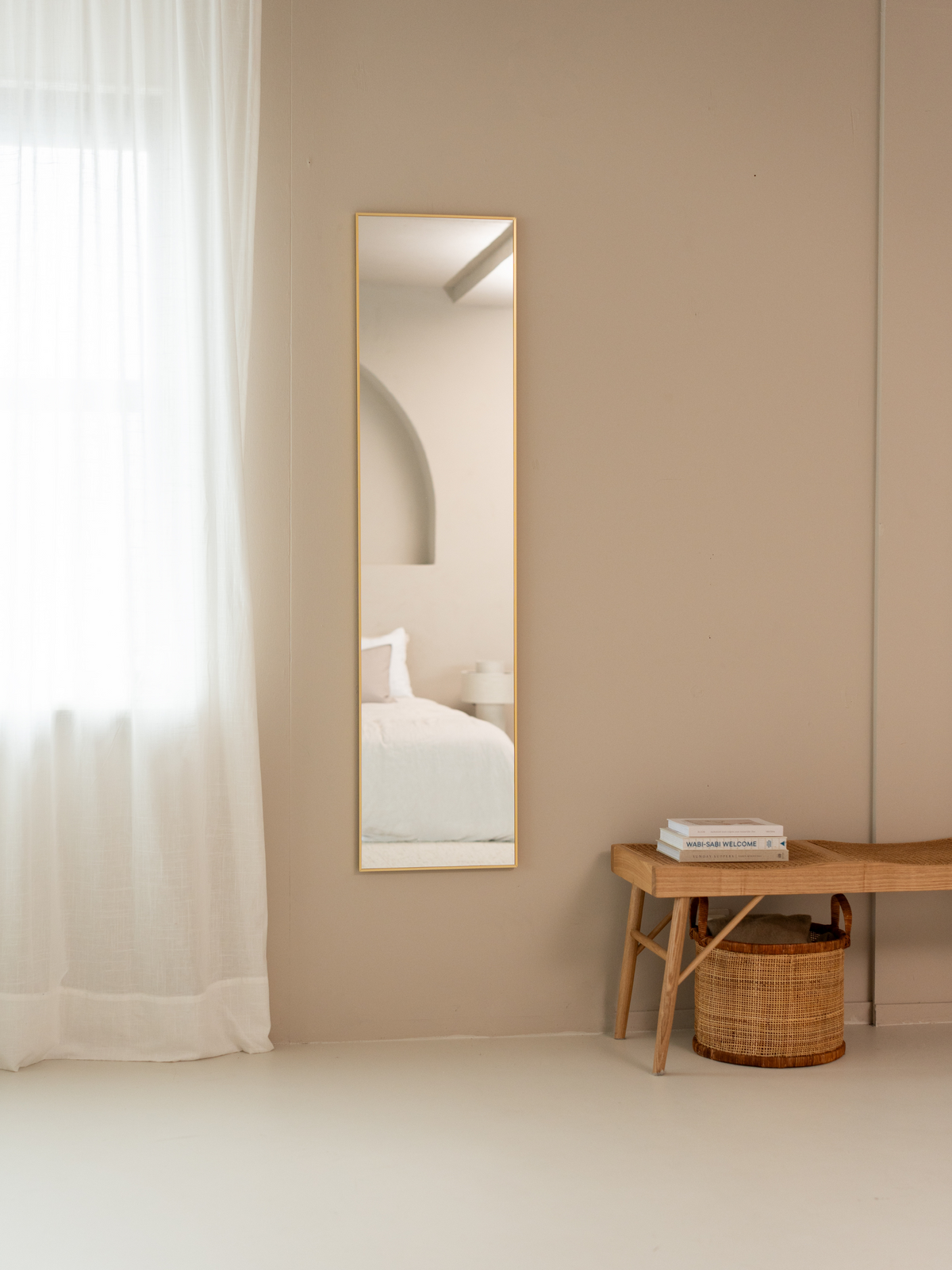 Tall rectangular Angelo mirror with slim gold frame hung on warm beige bedroom wall beside sheer white curtain and rattan…