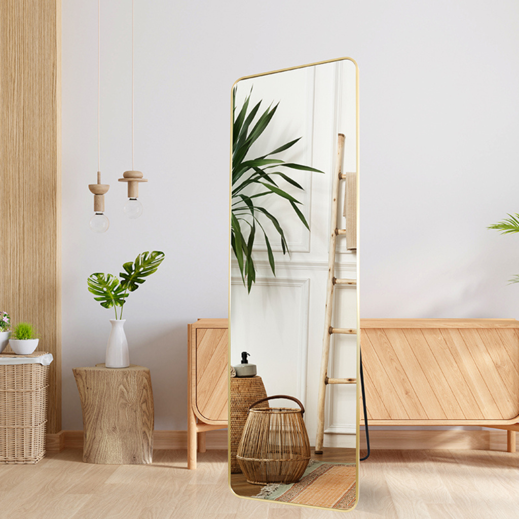 Gold full-length mirror with rounded corners leaning against wall in boho living room, reflecting palm plant and rattan ba…