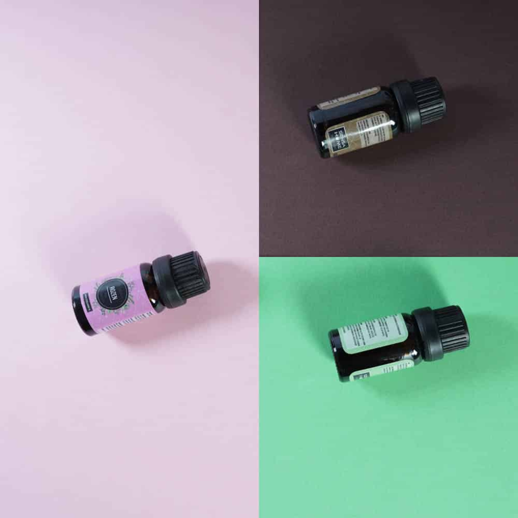 Three dark amber 10ml essential oil bottles lying on their sides across split pink, brown, and green backgrounds, overhead…
