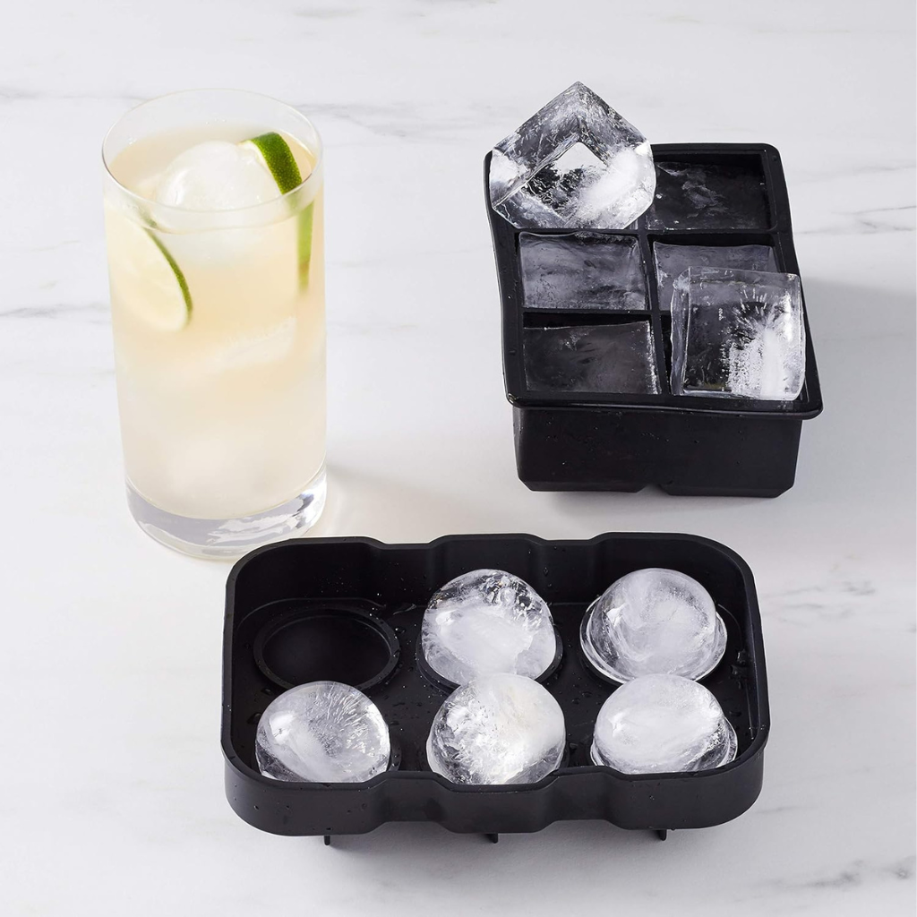 Two black silicone ice trays on marble surface; front tray holds 5 round ice spheres, back tray holds large square cubes,…