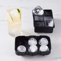 Two black silicone ice trays on marble surface; front tray holds 5 round ice spheres, back tray holds large square cubes,…