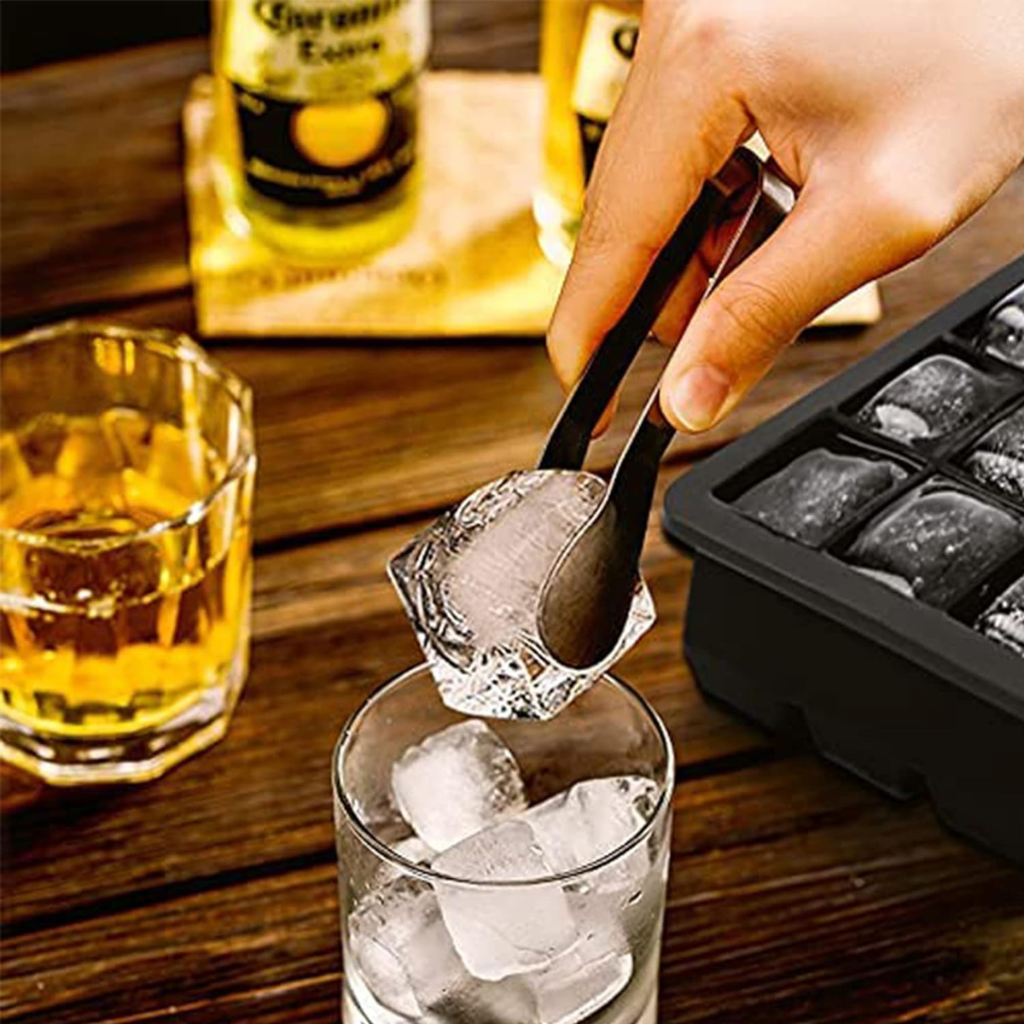 Hand using black tongs to transfer a large ice cube from a black silicone tray into a glass, whisky and beer bottles in ba…