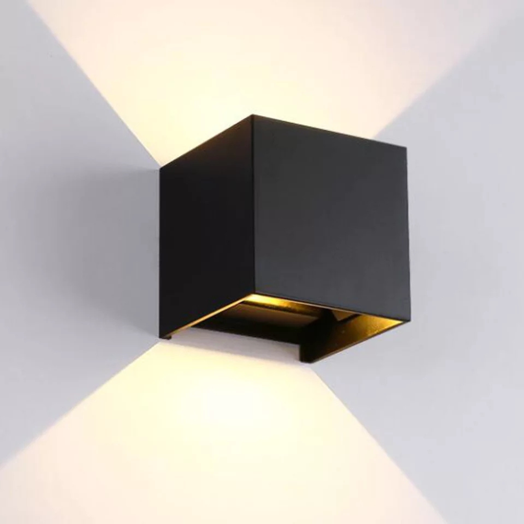 Black cube LED wall lamp mounted on grey wall, casting warm symmetrical up-down light beams, gold interior visible through…