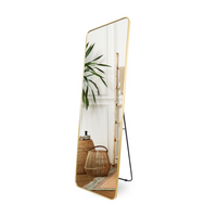 Gold full-length mirror with brushed aluminum frame standing on black metal easel, angled view, reflecting a boho-styled r…