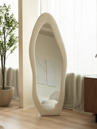Full-length asymmetric mirror with cream suede velvet frame leaning against wall in bright room with olive tree and sheer…