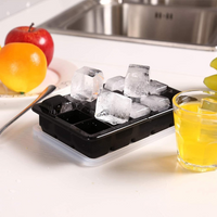 Black silicone ice cube tray filled with large clear cubes beside a glass of orange juice, fruit and kitchen sink in backg…