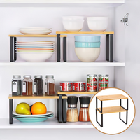 Lifestyle shot of two bamboo-top black metal cabinet risers organising bowls, plates, spice jars, and canned goods inside…