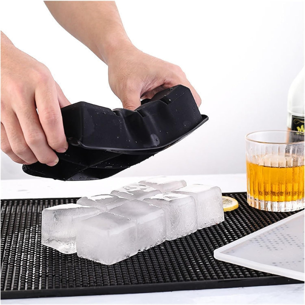 Hands peeling black silicone ice cube tray to release large clear ice cubes onto a bar mat, whisky glass alongside