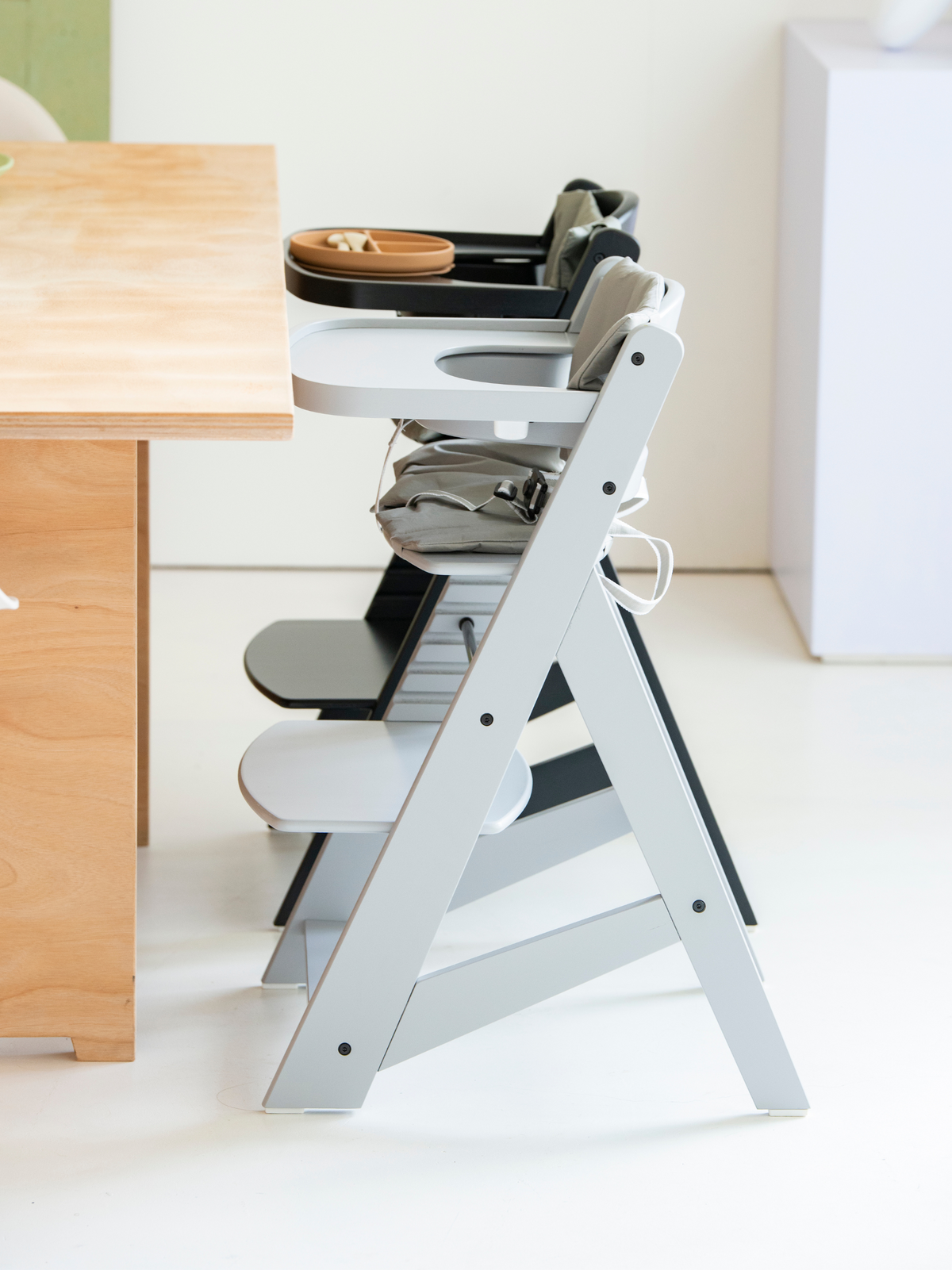 Grey wooden highchair side view beside a dining table, with tray holding a small plate, grey cushion, and adjustable footrest