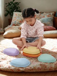 Young girl sitting cross-legged on a floral mat, stacking pastel balance stones; purple, blue, mint, orange, and yellow pi…