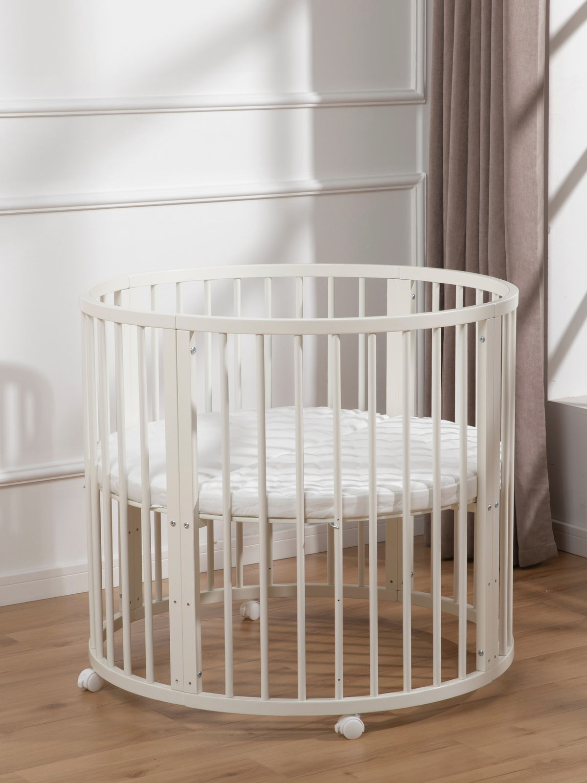 White round wooden baby playpen with mattress inside, angled view, lockable castors, beside curtain on wood floor