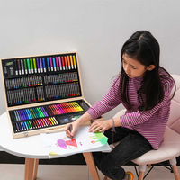 Girl in striped top drawing with markers at a table, open 180-piece wooden art set beside her showing rows of colourful su…
