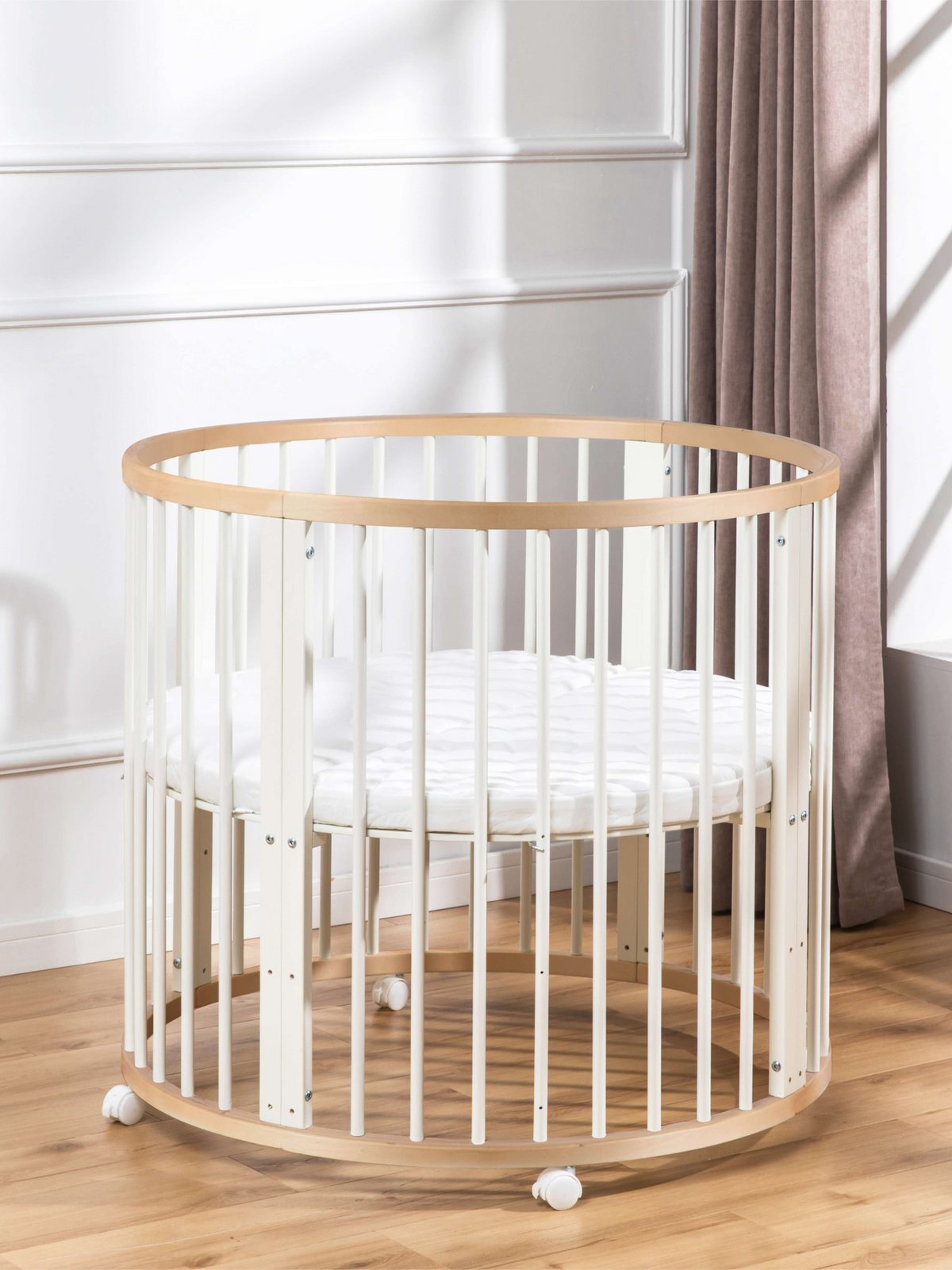 Round baby playpen with white slats and natural wood rim, shown with white quilted mattress inside, on castors, beside a c…