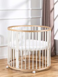 Round baby playpen with white slats and natural wood rim, shown with white quilted mattress inside, on castors, beside a c…