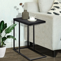 Black C-shaped side table tucked beside cream sofa, cup and vase on matte wood top, tropical plant and wood floor in backg…