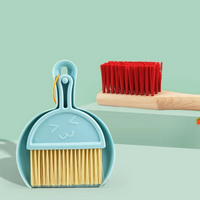 Blue mini plastic dustpan and brush set with smiley face detail, beside a red-bristled wooden scrub brush on a green surface