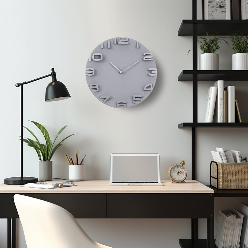Grey round wall clock with 3D raised numerals mounted above a modern home office desk with laptop and black shelving unit…