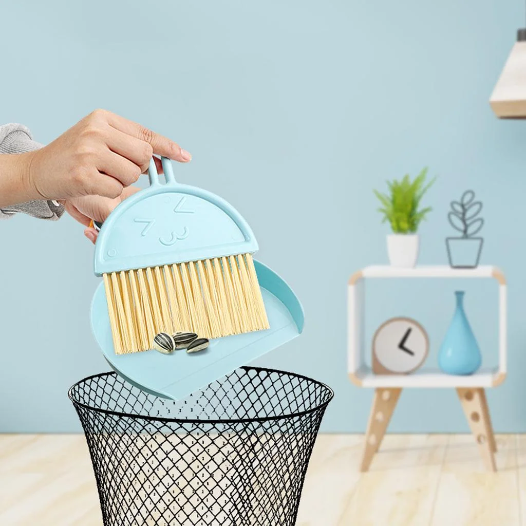 Hand tipping a light blue mini dustpan with cream bristle brush over a black mesh bin, emptying sunflower seeds indoors
