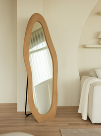 Taupe suede velvet asymmetric floor mirror leaning against cream wall beside white armchair, black metal stand visible at…
