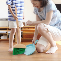 Woman crouching on wood floor holding light blue dustpan while child sweeps with green-bristled wooden broom indoors
