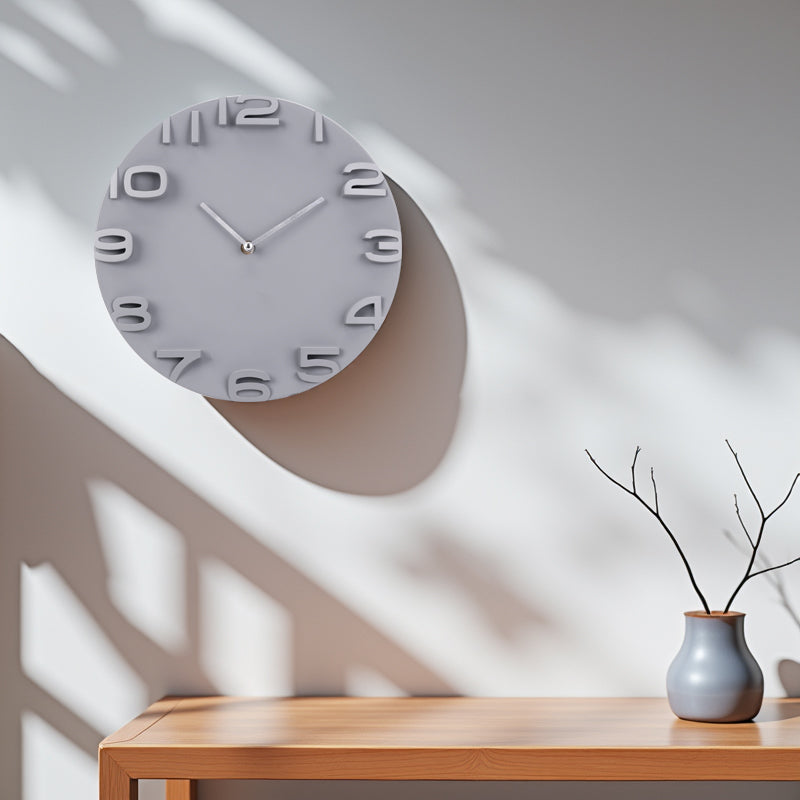 Grey round wall clock with 3D raised numerals hung on sunlit white wall above a wooden console table with a small vase