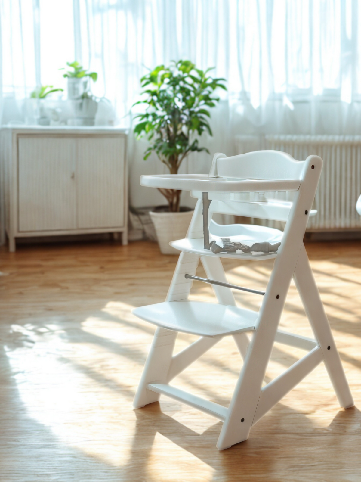 White wooden high chair with tray and grey safety harness, lifestyle shot on oak floor with plants and curtains in background