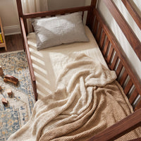 Overhead view of walnut brown wooden toddler bed with safety rails, cream fitted sheet, knit blanket, and grey pillow in s…