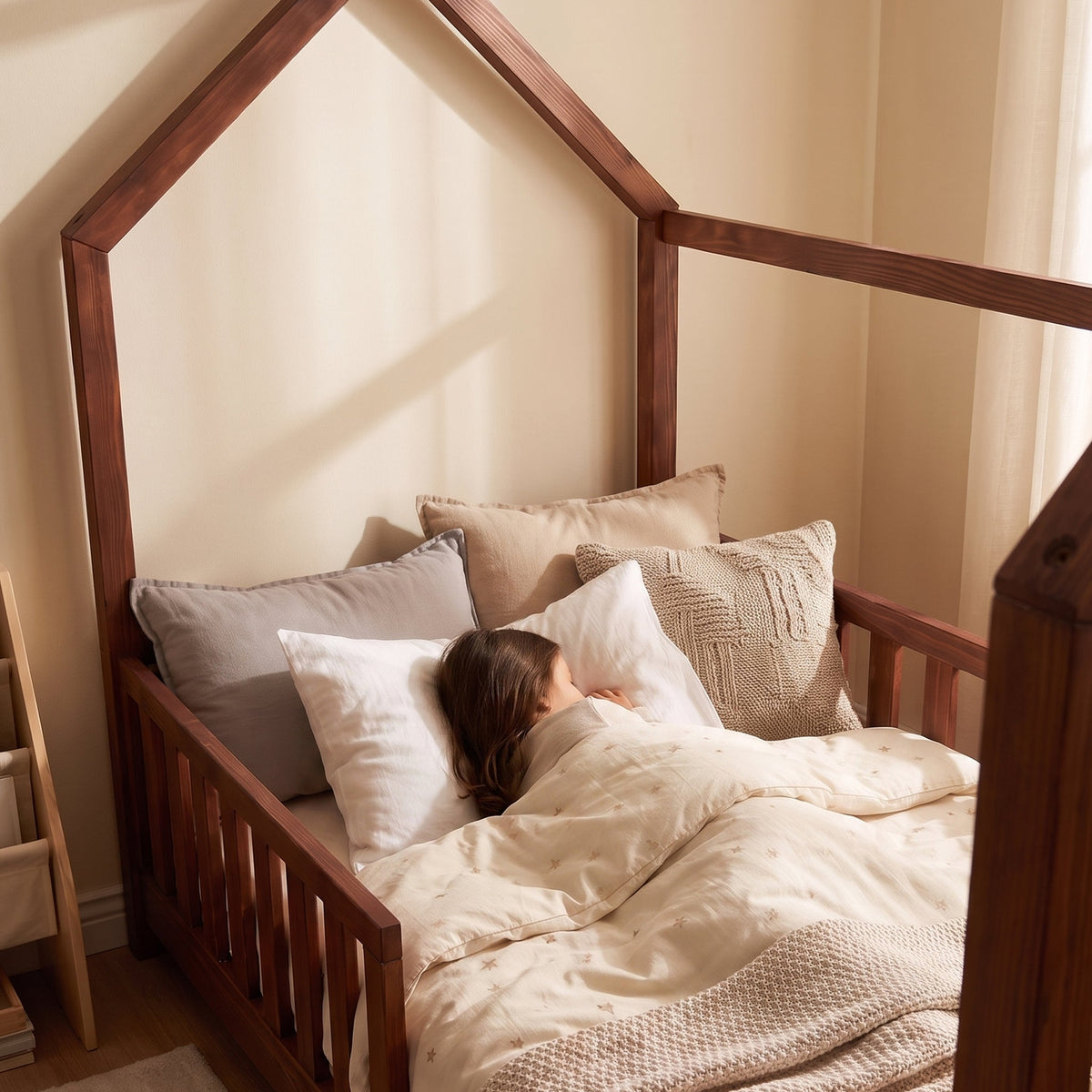 Child sleeping in walnut brown wooden house-frame bed with safety rails, cream star-print duvet and mixed neutral cushions…