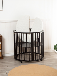 Black round wooden playpen with mattress propped upright inside, adjustable base visible, nursery setting with jute rug an…