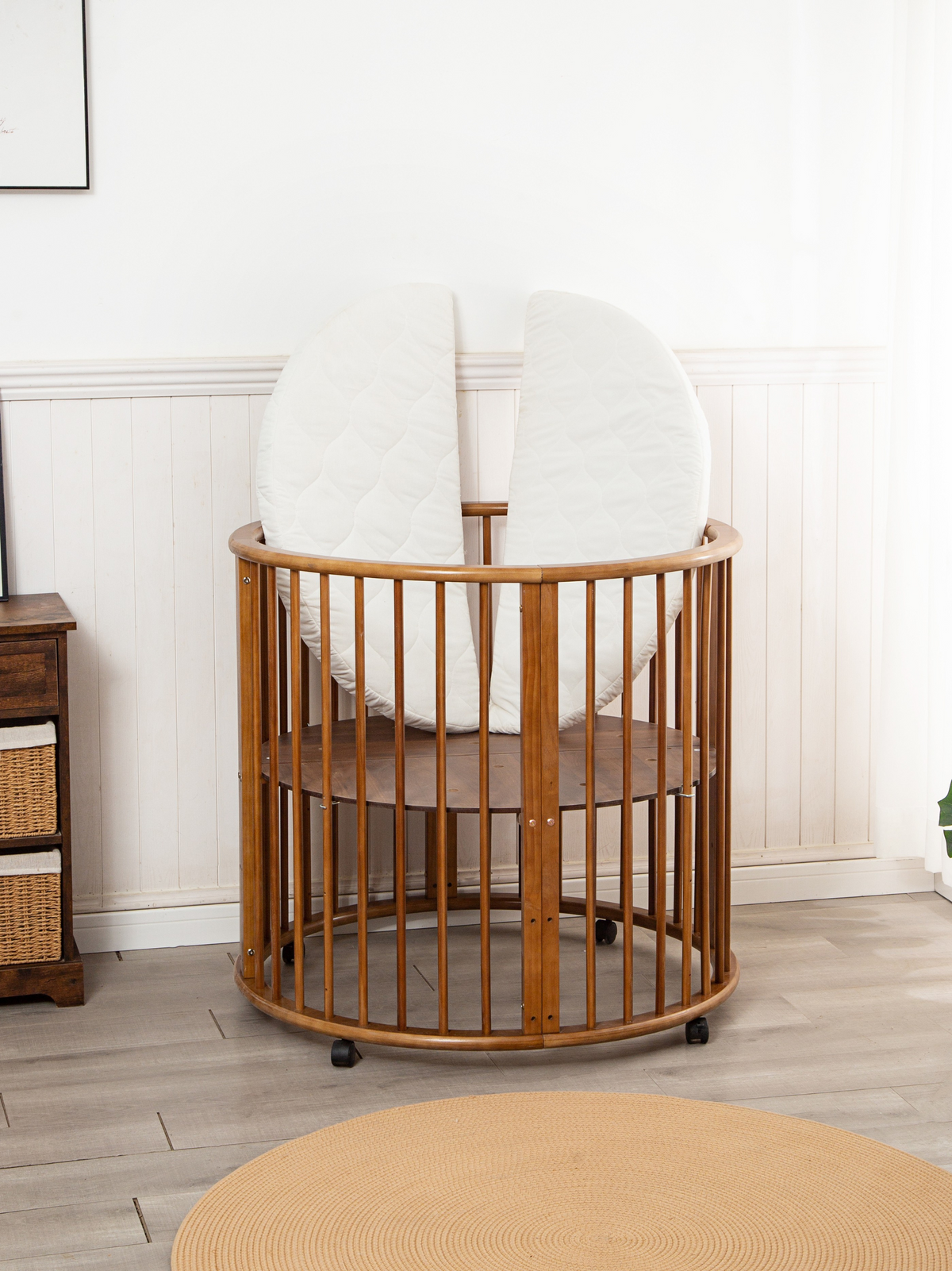 Brown round wooden baby playpen on castors with two white half-circle mattress sections propped upright inside, nursery se…