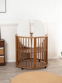 Brown round wooden baby playpen on castors with two white half-circle mattress sections propped upright inside, nursery se…