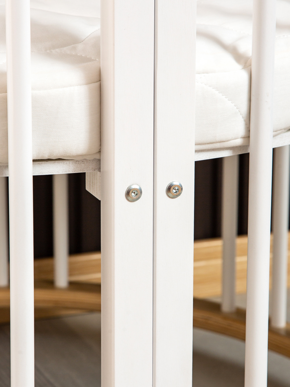Close-up of two silver hex bolts on white wooden slats joining the round playpen frame, with white mattress and wood floor…