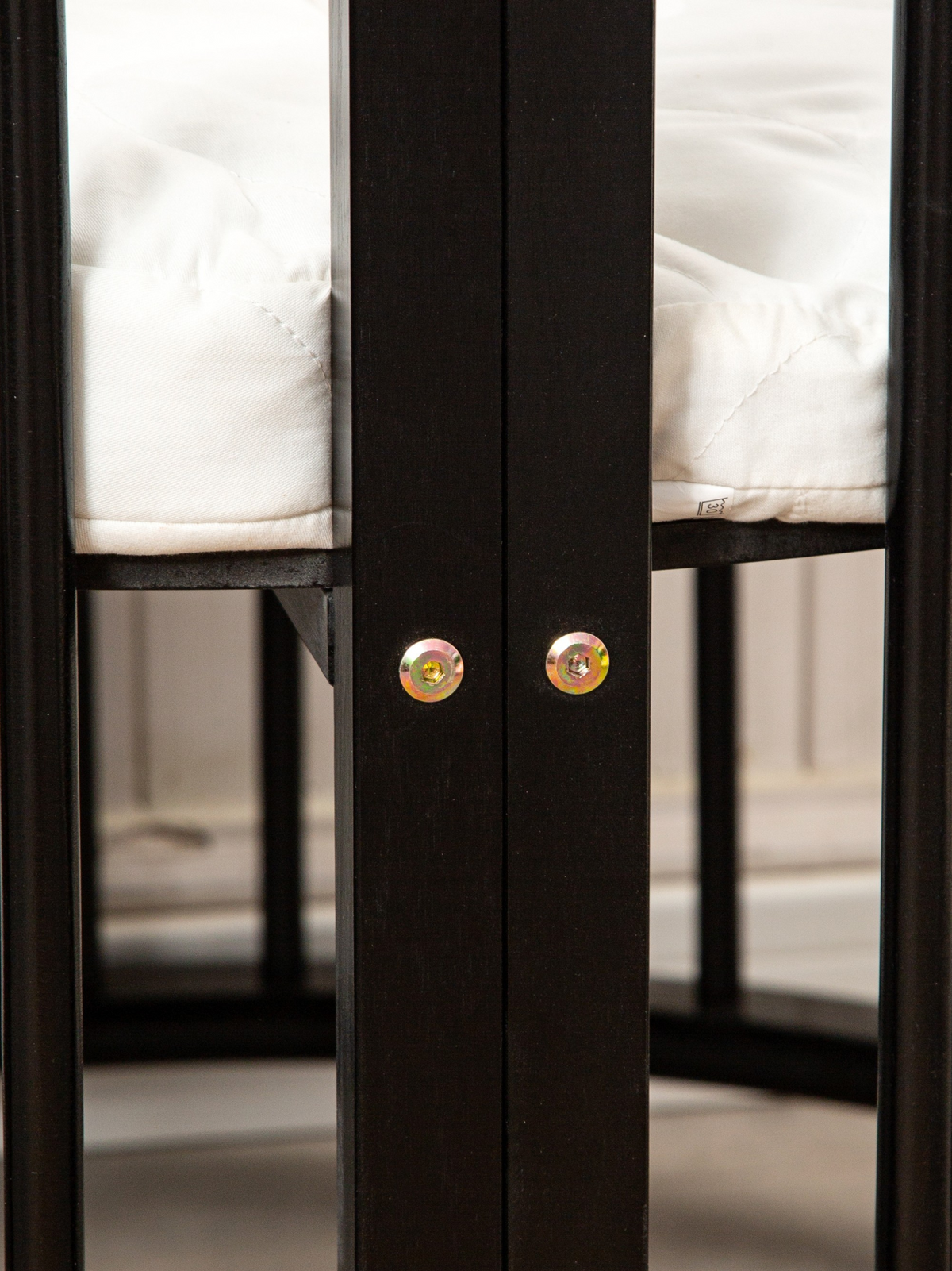 Close-up of two gold screws on a black wooden playpen post, with white quilted mattress and slatted frame visible behind