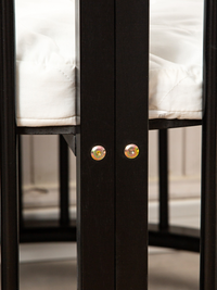 Close-up of two gold screws on a black wooden playpen post, with white quilted mattress and slatted frame visible behind
