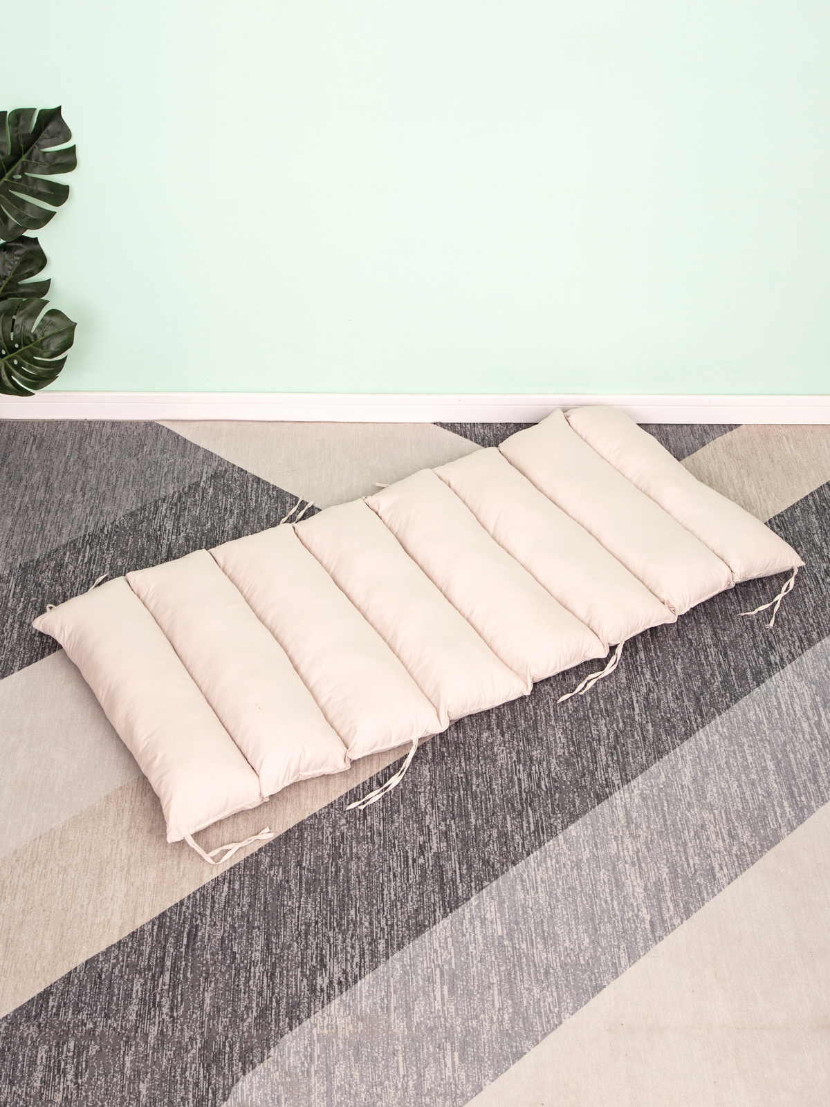 Cream segmented floor cushion with tie strings laid flat on grey striped carpet, monstera plant and mint wall in background