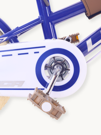 Close-up of white chain guard with blue circle detail, brown plastic pedal, and chrome crank on dark blue children's bike…