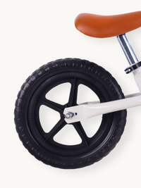 Close-up of rear wheel on white balance bike, showing black rubber knobbed tire, five-spoke plastic rim, and tan saddle above