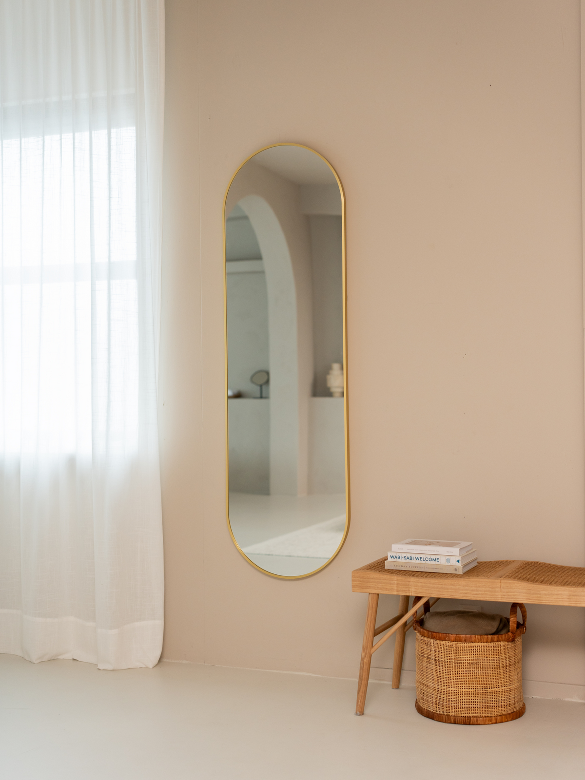 Tall gold capsule mirror on beige wall beside sheer white curtain, reflecting arched doorway, with rattan bench and basket…