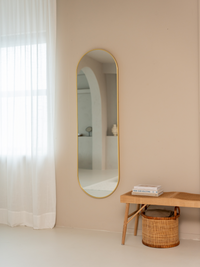 Tall gold capsule mirror on beige wall beside sheer white curtain, reflecting arched doorway, with rattan bench and basket…