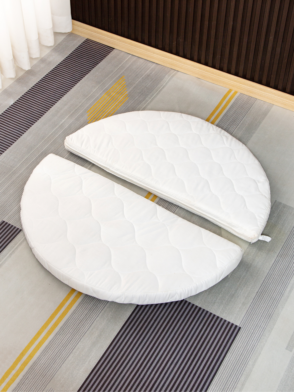 Two-piece white quilted circular mattress split in half, laid flat on a striped rug beside a wooden furniture base, overhe…