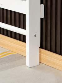 Close-up of white wooden climbing frame post base showing bolt fixing, against oak skirting board and dark ribbed wall panel
