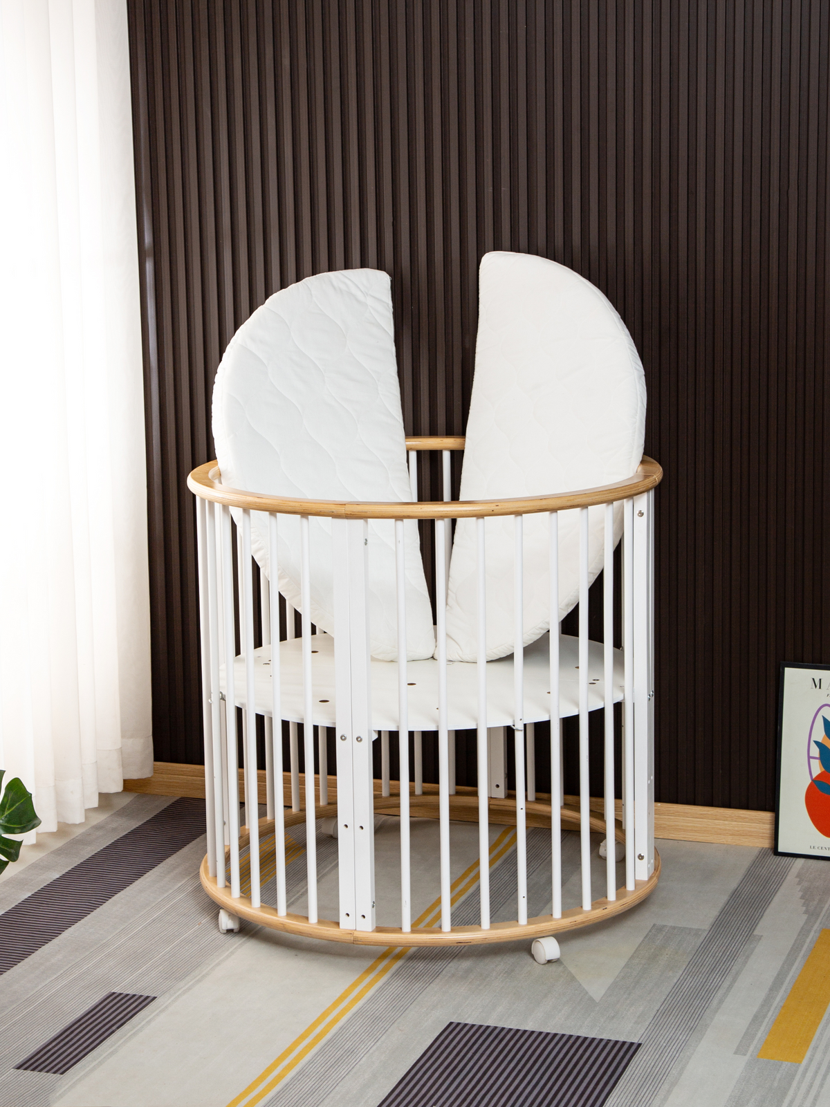 Round wooden playpen with white slats and natural wood rim, two white quilted half-circle mattress sections propped uprigh…