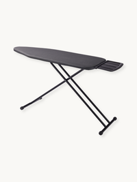 Black ironing board angled side view showing dark grey cotton cover, iron rest holder at wide end, and black cross-leg ste…