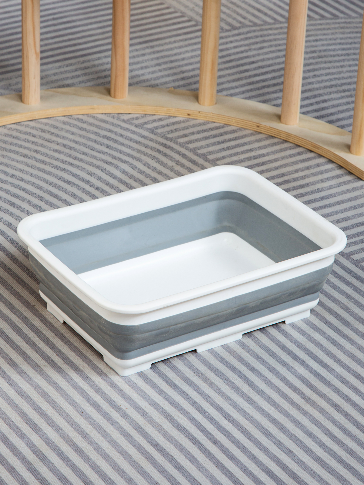 Grey and white collapsible square storage bin shown removed from climbing arch, placed on grey striped rug with wooden fra…