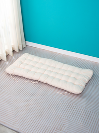 Cream tufted rectangular cushion with tie fasteners laid flat on patterned carpet against teal wall, overhead angle