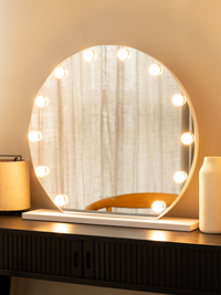 Round white Hollywood vanity mirror with warm-toned bulbs glowing, styled on a dark wood dressing table beside a lamp and…