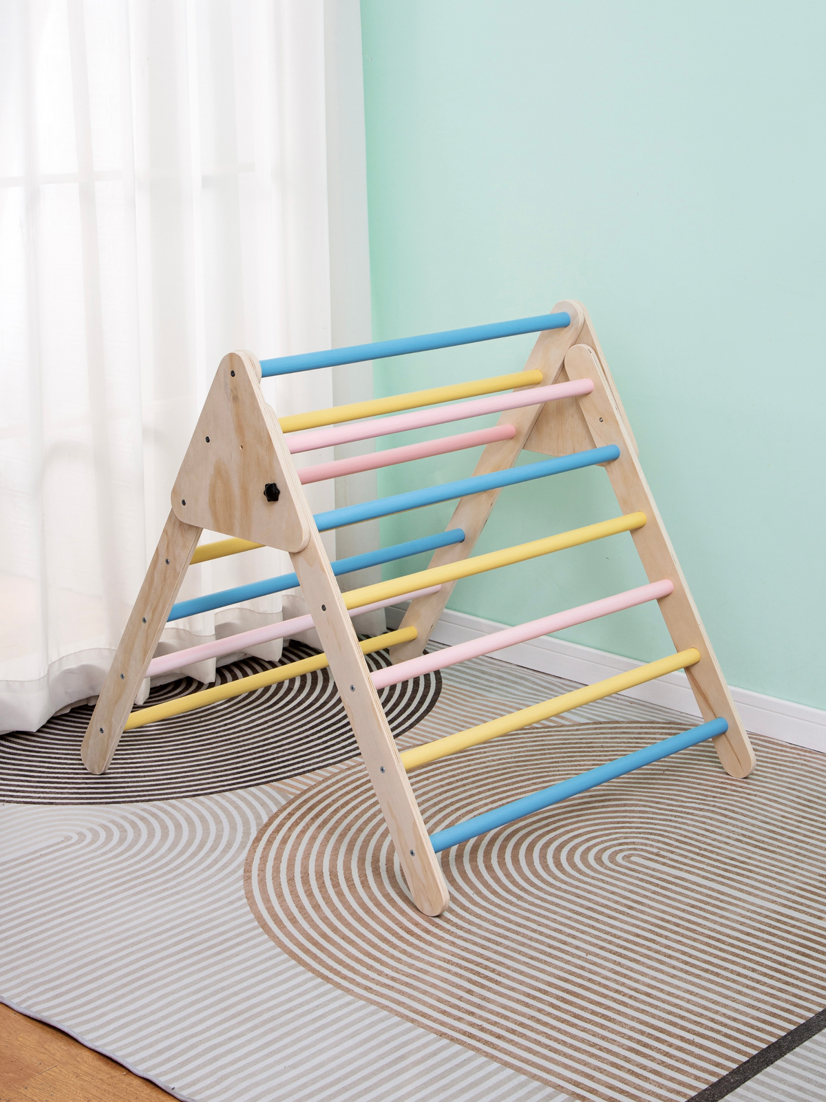 Wooden Pikler triangle climbing frame with blue, pink and yellow rungs, three-quarter view on a geometric rug near a window