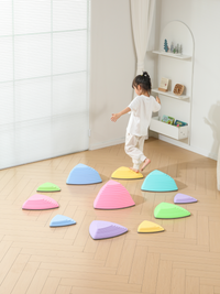 Young girl stepping across pastel triangular balance stones on herringbone floor indoors, stones in multiple sizes and col…