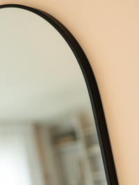 Close-up of slim matte black aluminium frame on arched top of oval mirror, mounted on peach-toned wall